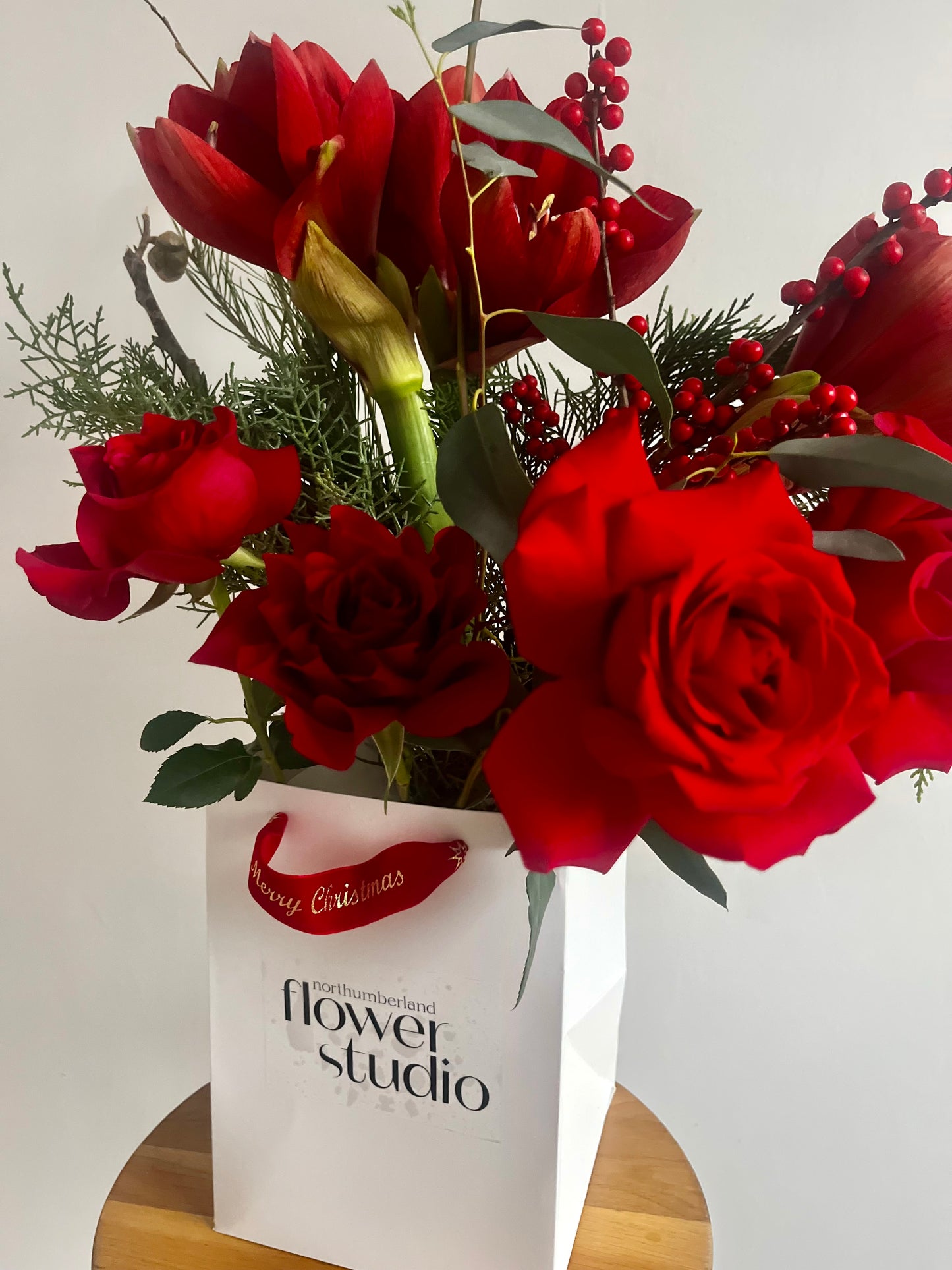 Bouquet of red Christmas flowers in a white bag with 'Northumberland Flower Studio' branding on a light background.