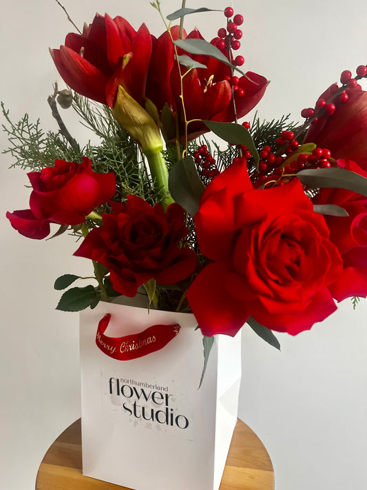 Bouquet of red Christmas flowers in a white bag with 'Northumberland Flower Studio' branding on a light background.
