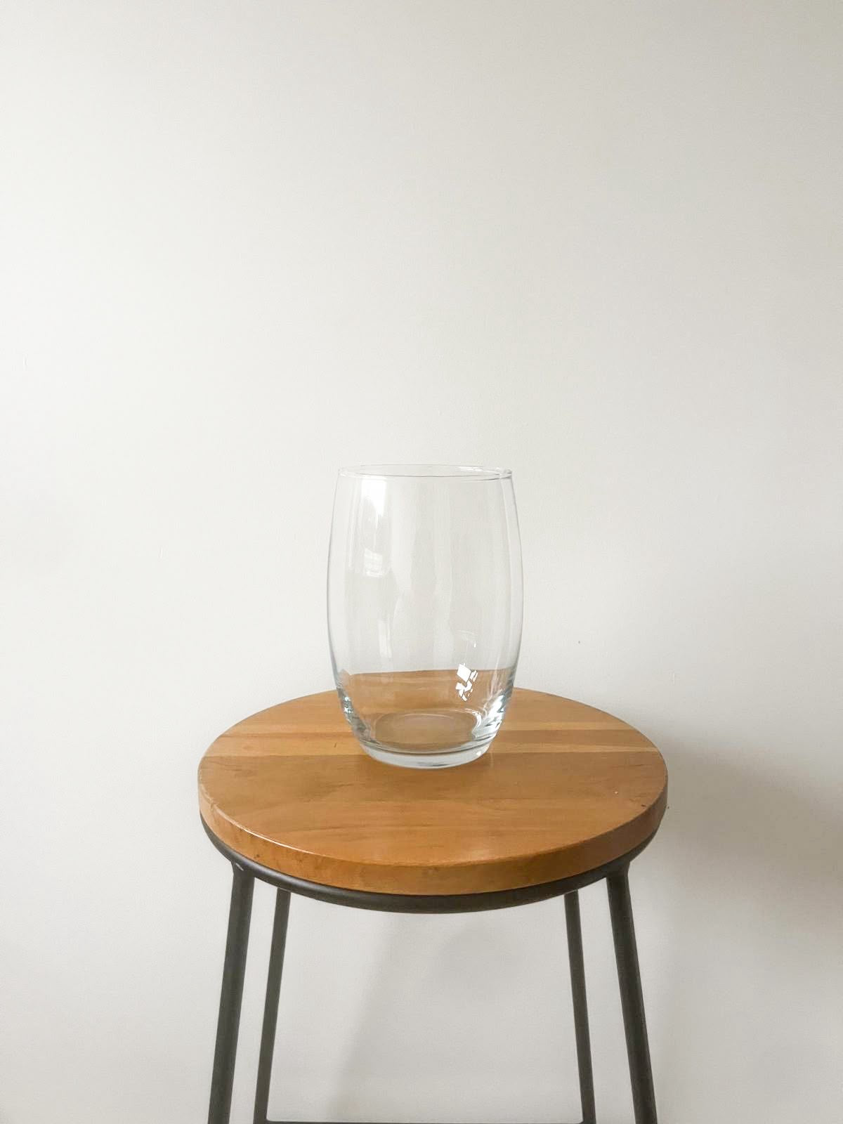 A clear glass tumbler with a flared rim sits centered on a round wooden stool with black metal legs against a plain off-white wall.