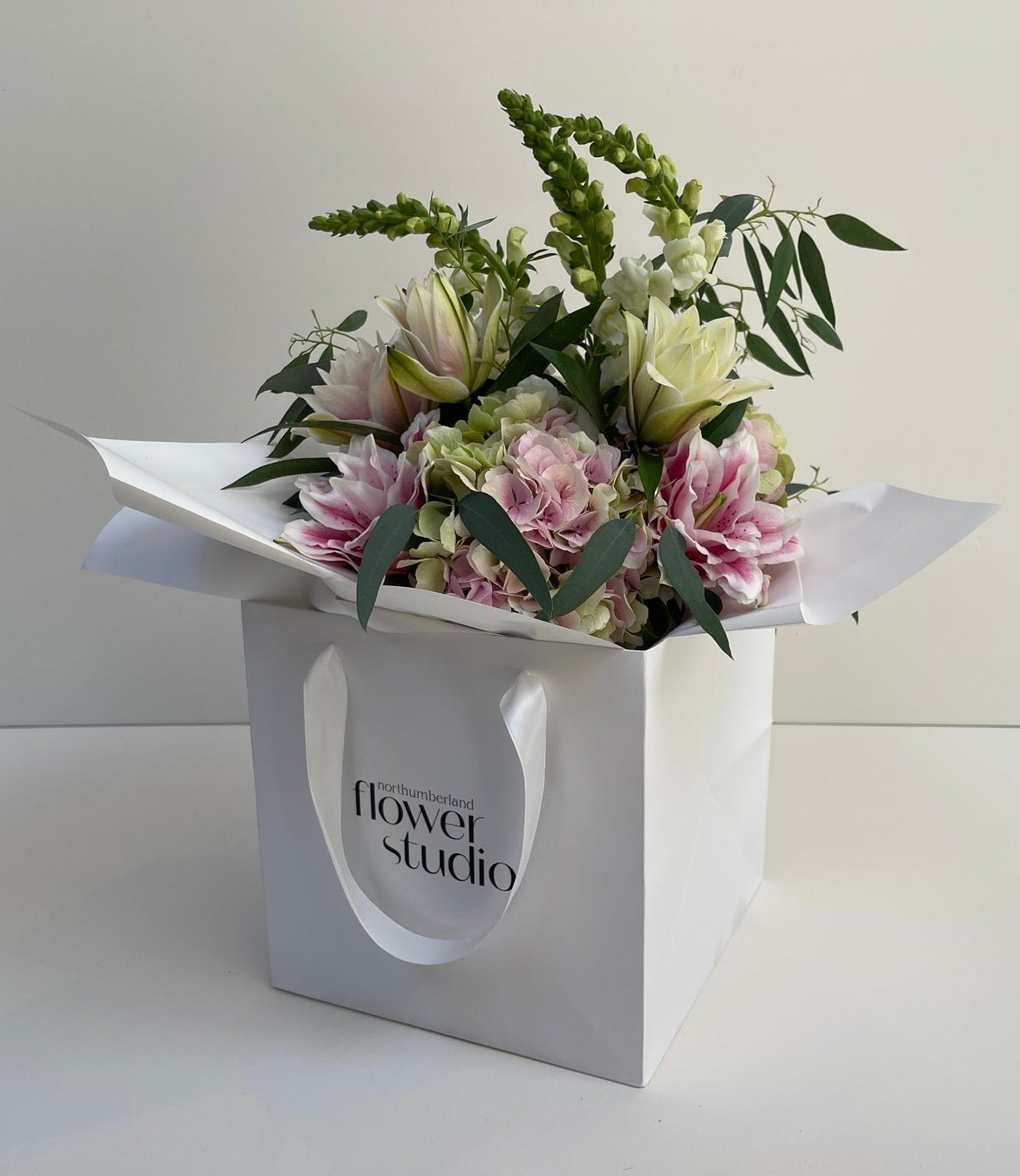 Hand-tied floral arrangement in a white gift bag with 'Northumberland Flower Studio' branding on a light background