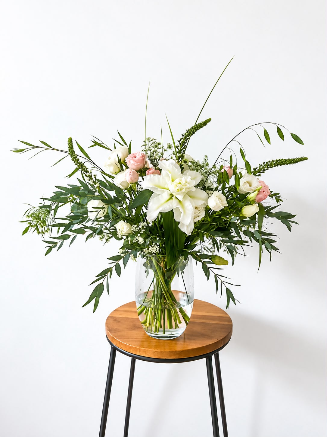 A fresh floral arrangement in a clear glass vase, featuring a large white lily, pink roses, and green foliage, displayed on a round wooden stool with black metal legs against a plain white wall.