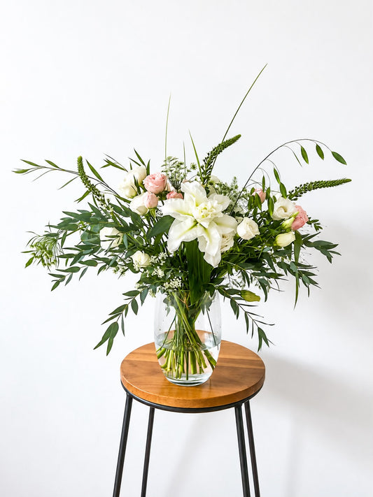 A fresh floral arrangement in a clear glass vase, featuring a large white lily, pink roses, and green foliage, displayed on a round wooden stool with black metal legs against a plain white wall.